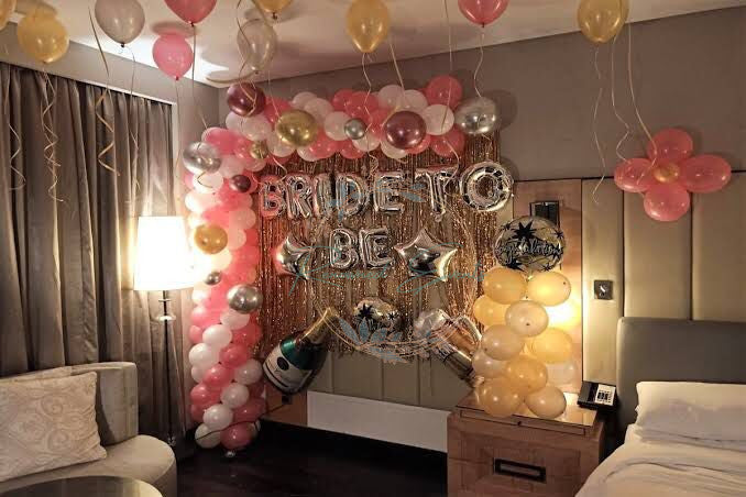 Rose-gold “BRIDE TO BE” balloon letters suspended in front of a glittering gold fringe curtain, flanked by clusters of blush pink, white, and metallic gold balloons, with oversized champagne bottle and diamond-ring balloons adding playful flair to a chic hotel-suite celebration.