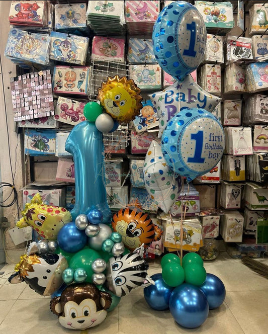 balloon arrangement for a baby boy's first birthday, featuring a large blue number "1" balloon surrounded by metallic animal-shaped balloons including a lion, monkey, zebra, and giraffe. The display also includes blue and silver balloons, along with round and star-shaped foil balloons reading "Baby Boy" and "1st Birthday." In the background, shelves are stocked with colorful party supplies and themed decorations.