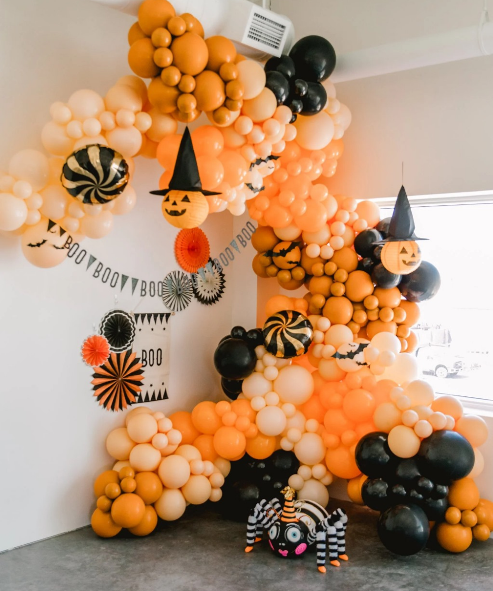 Halloween balloon wall with orange and black balloons, pumpkin faces with witch hats, spider prop and festive décor for indoor party setup.