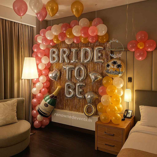 A hotel room decked out for a bachelorette party: a gold tinsel backdrop with silver “BRIDE TO BE” balloon letters, surrounded by pink, white, and gold balloon clusters, plus oversized champagne bottle and diamond-ring balloons.