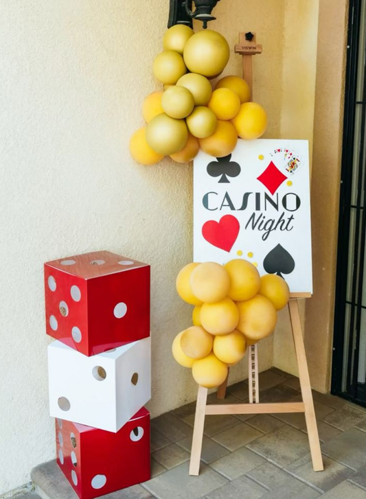 Casino Night party entrance decoration with stacked red and white dice props, golden balloon clusters, and a playing card themed welcome board.