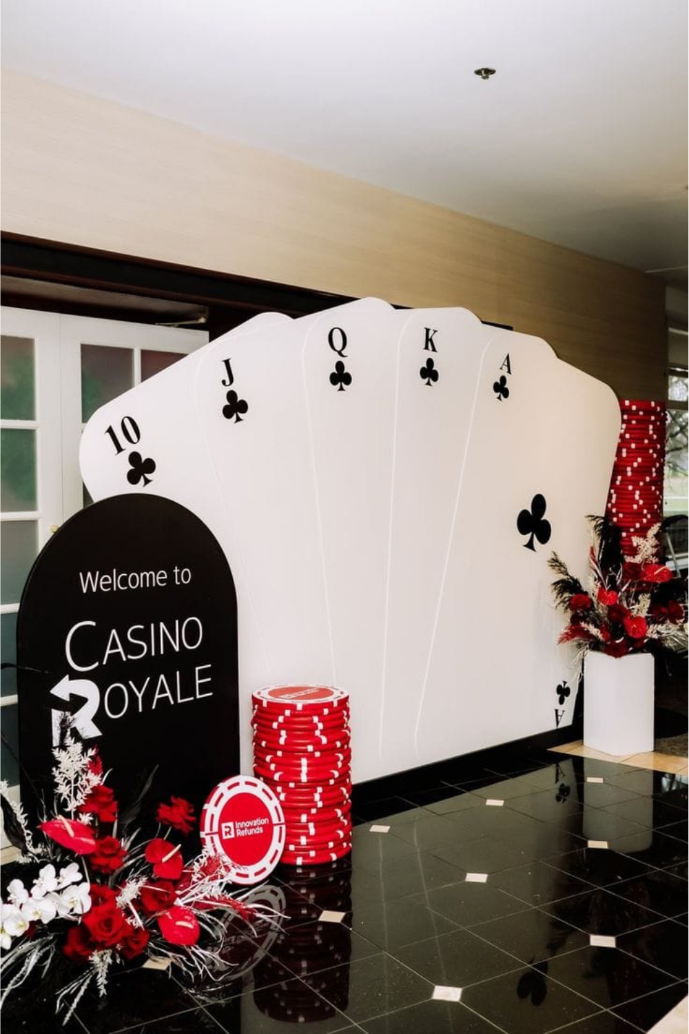 Elegant Casino Royale backdrop decoration featuring giant playing cards, red poker chips, and floral arrangements in black, red, and white theme — perfect for casino night parties, birthdays, and corporate events.