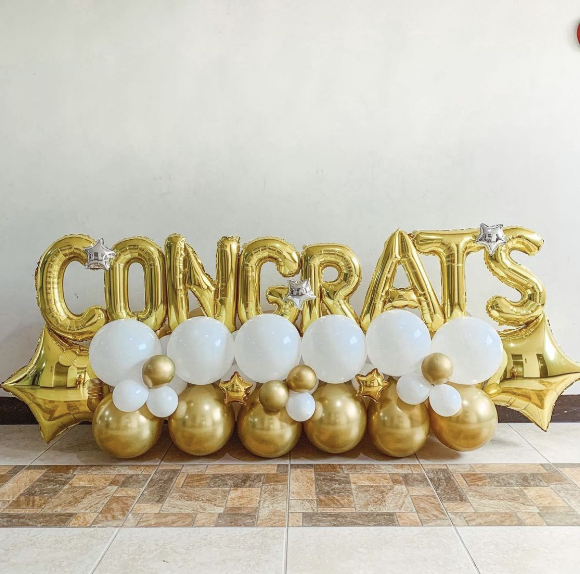 Gold “CONGRATS” foil balloon letters with white and gold balloon clusters arranged at the base, decorated with small star balloons.