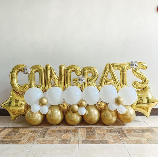 Gold “CONGRATS” foil balloon letters with white and gold balloon clusters arranged at the base, decorated with small star balloons.