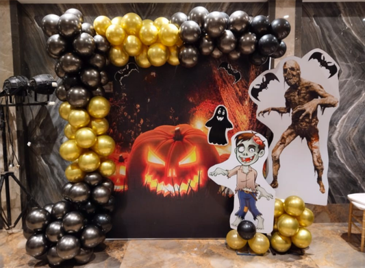 Halloween backdrop with black and golden balloon arch, zombie cutout, ghost prop and glowing pumpkin design.