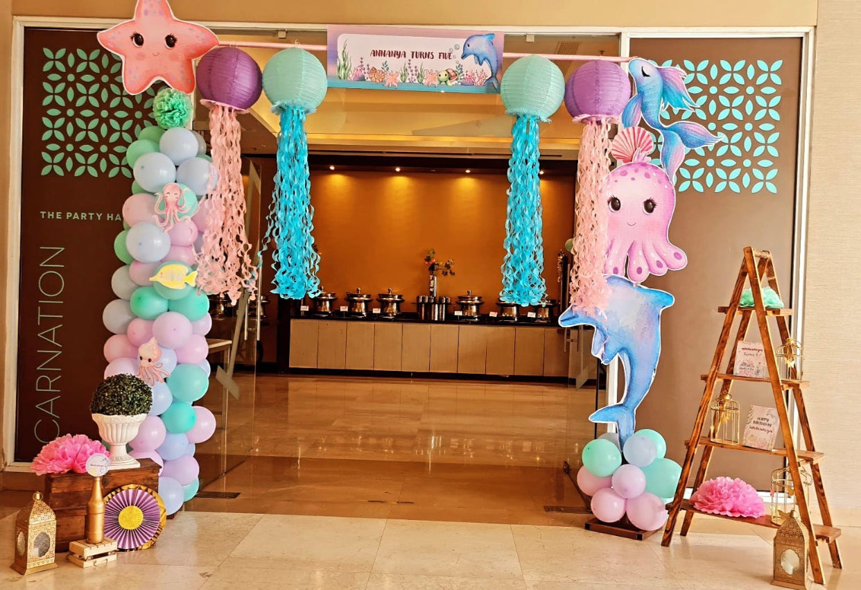 Kids ocean theme party entrance decorated with pastel balloons, jellyfish lanterns, starfish, octopus, and dolphin props.