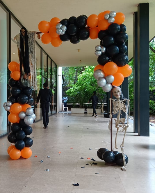 Halloween entrance decoration featuring a spooky skeleton prop with black, orange, and silver balloon arch.